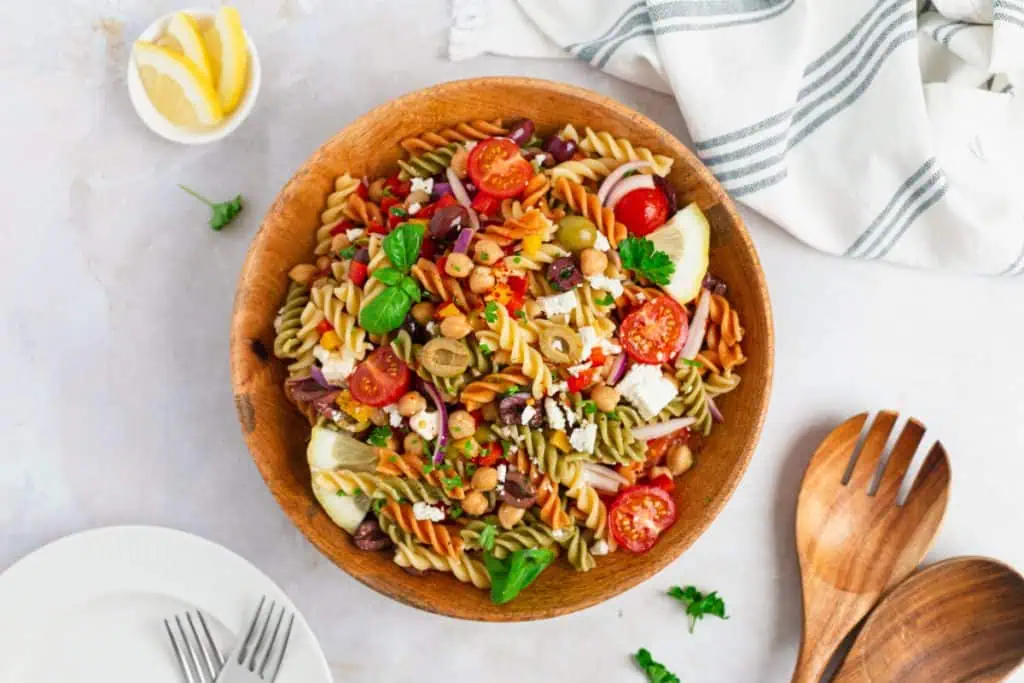 Mediterranean pasta salad in a brown bowl with feta crumbles, halved olives, colorful peppers, and cherry tomatoes in honey lemon dressing, wooden spoons on a white marble background