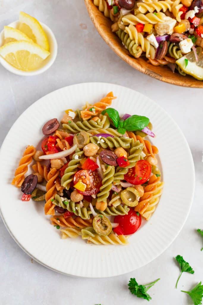 Mediterranean pasta salad in a large white plate with feta crumbles, halved olives, colorful peppers, and cherry tomatoes in honey lemon dressing
