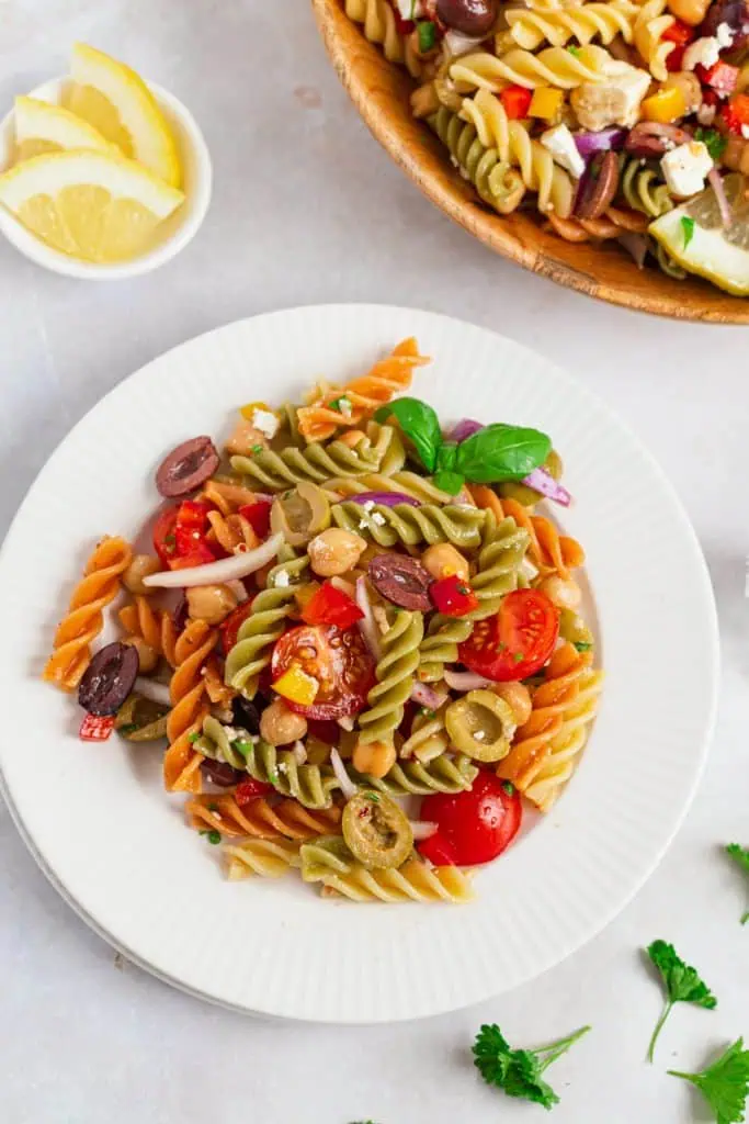 Mediterranean pasta salad in a large white plate with feta crumbles, halved olives, colorful peppers, and cherry tomatoes in honey lemon dressing