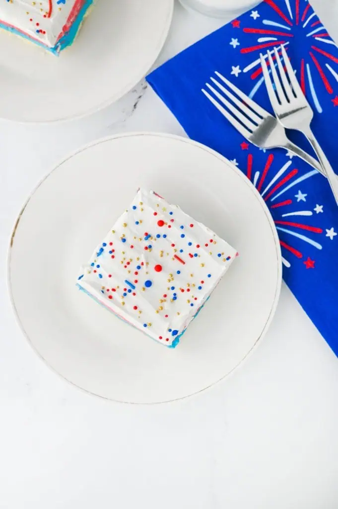 Red white and blue lush square on a white plate showing golden shortbread crust, blue cream cheese, red pudding, and Cool Whip layers with patriotic sprinkles shot overhead with patriotic napkin and forks