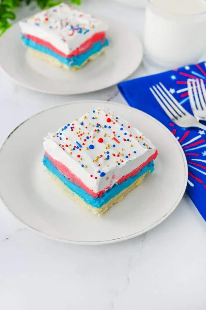 2 slices of Red white and blue lush square on a white plate showing golden shortbread crust, blue cream cheese, red pudding, and Cool Whip layers with patriotic sprinkles