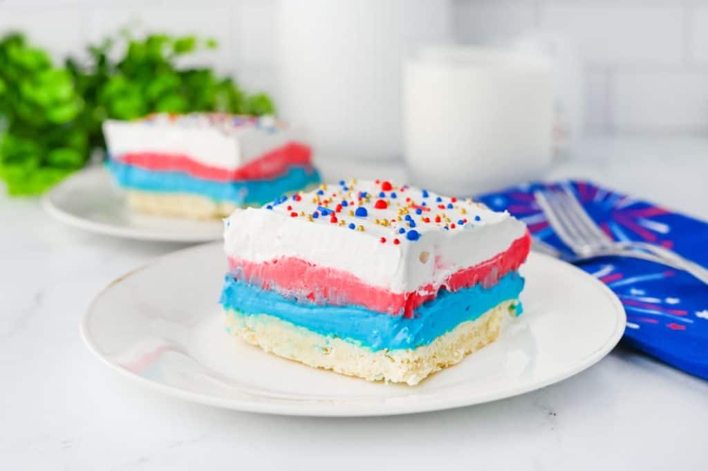 Close-up of 2 slices of red white and blue lush layers with crisp stripes of blue cheesecake, red pudding, and fluffy white whipped topping on shortbread crust