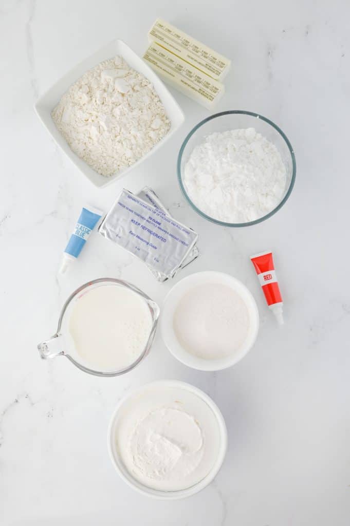 Red white and blue lush ingredients arranged overhead with flour, butter, cream cheese, cheesecake pudding mixes, milk, Cool Whip, and gel food coloring
