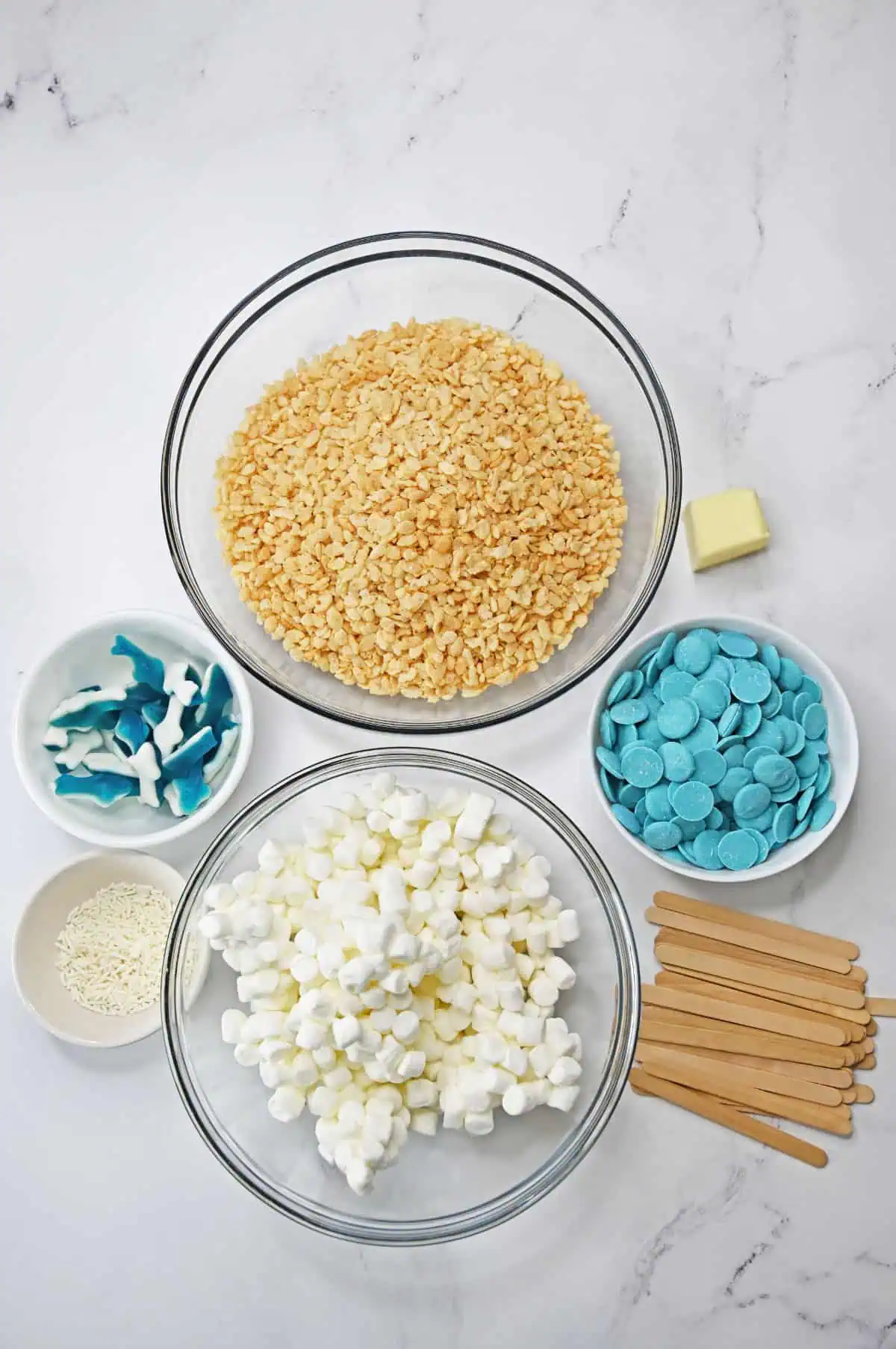 Overhead flatlay of shark rice krispie treat ingredients with marshmallows, rice cereal, blue candy melts, gummy sharks, and popsicle sticks