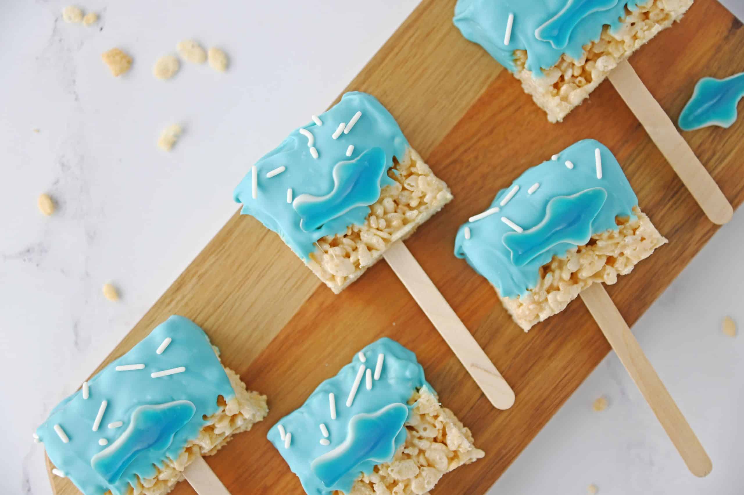 ) Close-up of a single shark rice krispie pop showing the chewy cereal, glossy blue candy coating, and a gummy shark on top