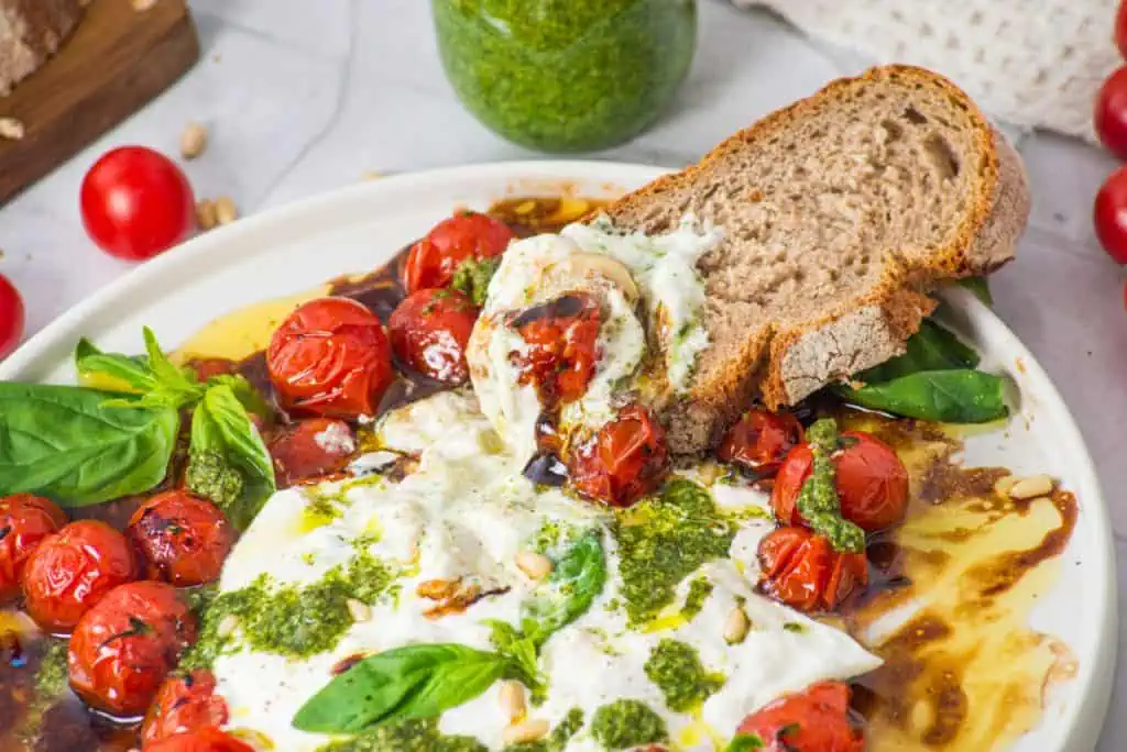 Tomato burrata salad platter served with crusty bread slices, a jar of homemade pesto, and fresh basil on a marble counter