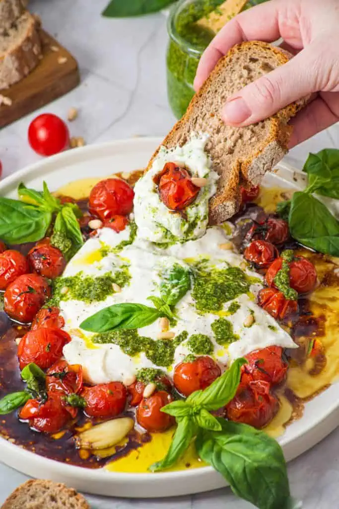 Tomato burrata salad platter served with a hand dipping crusty bread slices, a jar of homemade pesto, and fresh basil on a marble counter