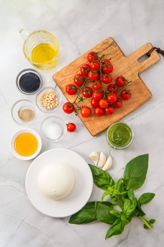 Tomato burrata salad ingredients arranged overhead with cherry tomatoes on the vine, burrata, pesto, honey, balsamic vinegar, garlic, basil, and pine nuts