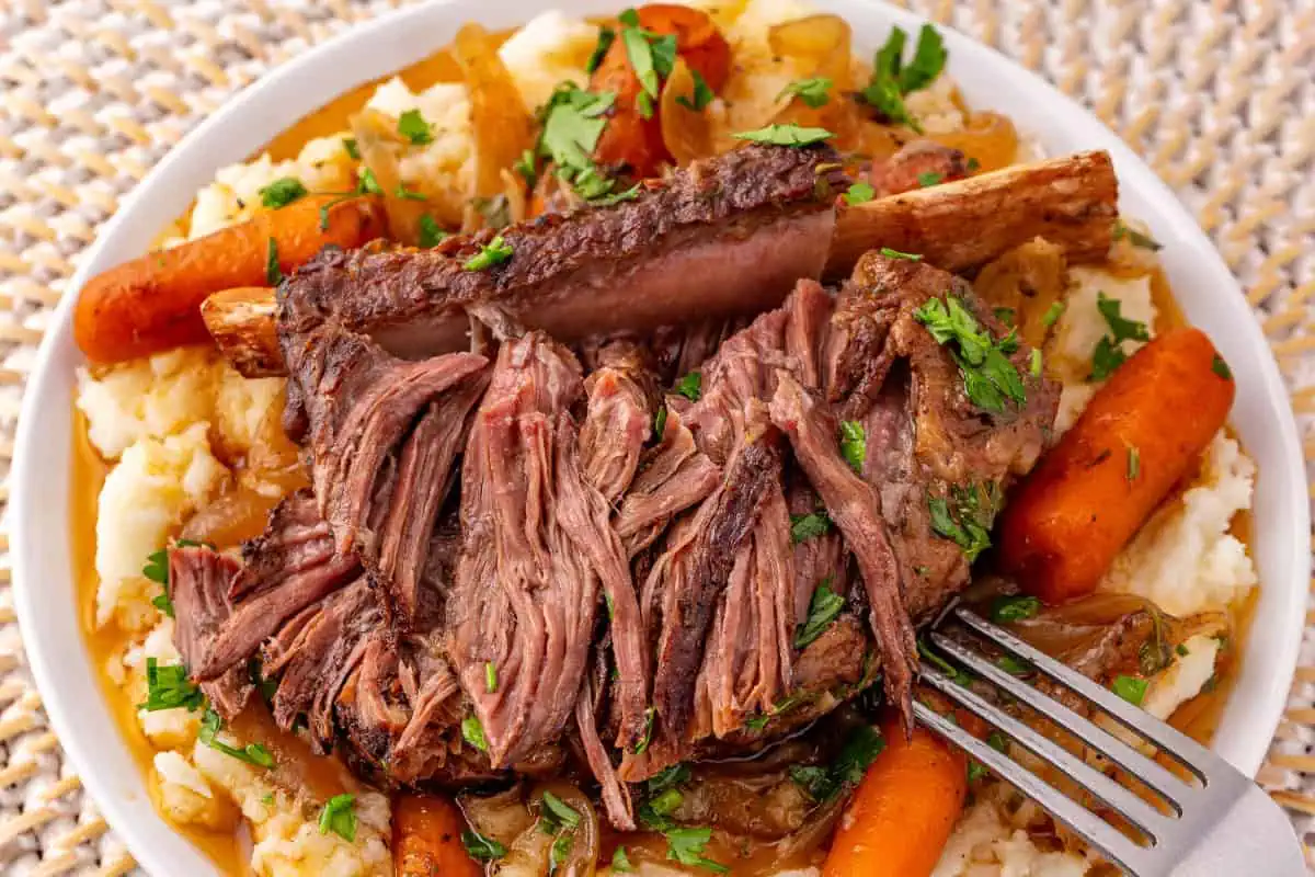 Close-up of fall-off-the-bone beef short ribs pulling apart with a fork in rich gravy with baby carrots over creamy mashed potatoes