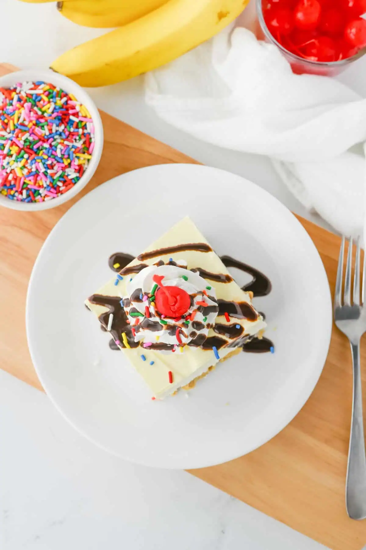 Banana Split Lush served on a dessert plate with whipped topping, fudge sauce, rainbow sprinkles, and a maraschino cherry.