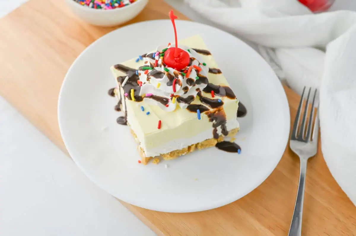 Sliced banana split lush square topped with whipped cream, fudge drizzle, rainbow sprinkles, and a maraschino cherry on a white plate