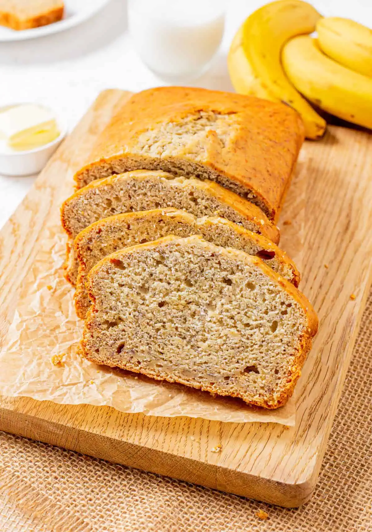 A golden loaf of sliced moist banana bread with a cracked top on a wooden cutting board with a ramekin of sliced butter tabs