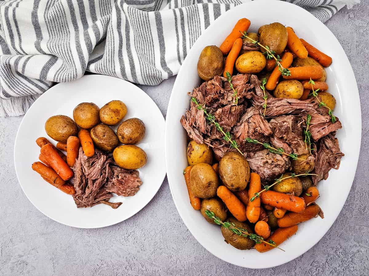 A plated serving of chuck roast with potatoes, carrots, and a spoonful of savory juices next the serving platter on white plates