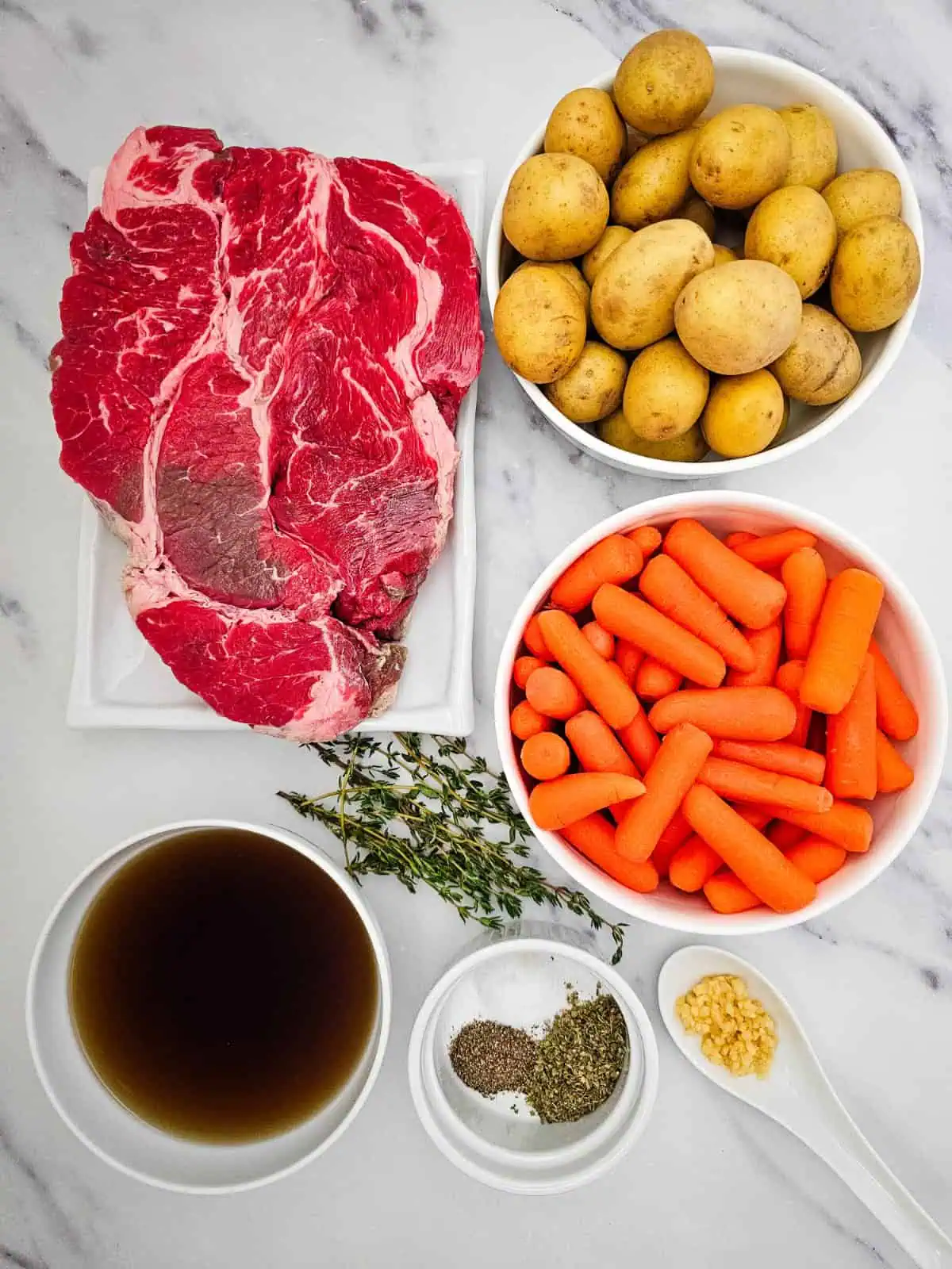 Chuck roast, baby potatoes, carrots, beef broth, garlic, oregano, and thyme arranged for slow cooker chuck roast