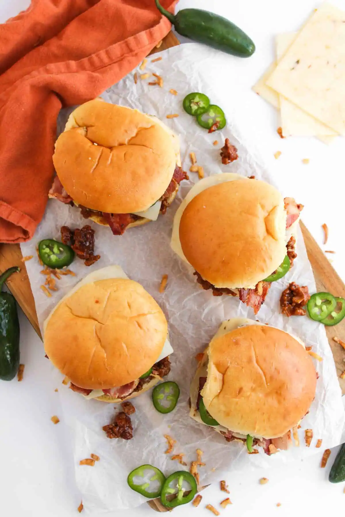 Four BBQ cowboy sloppy joes on a board with parchment paper surrounded by sliced jalapenos and crumbled bacon
