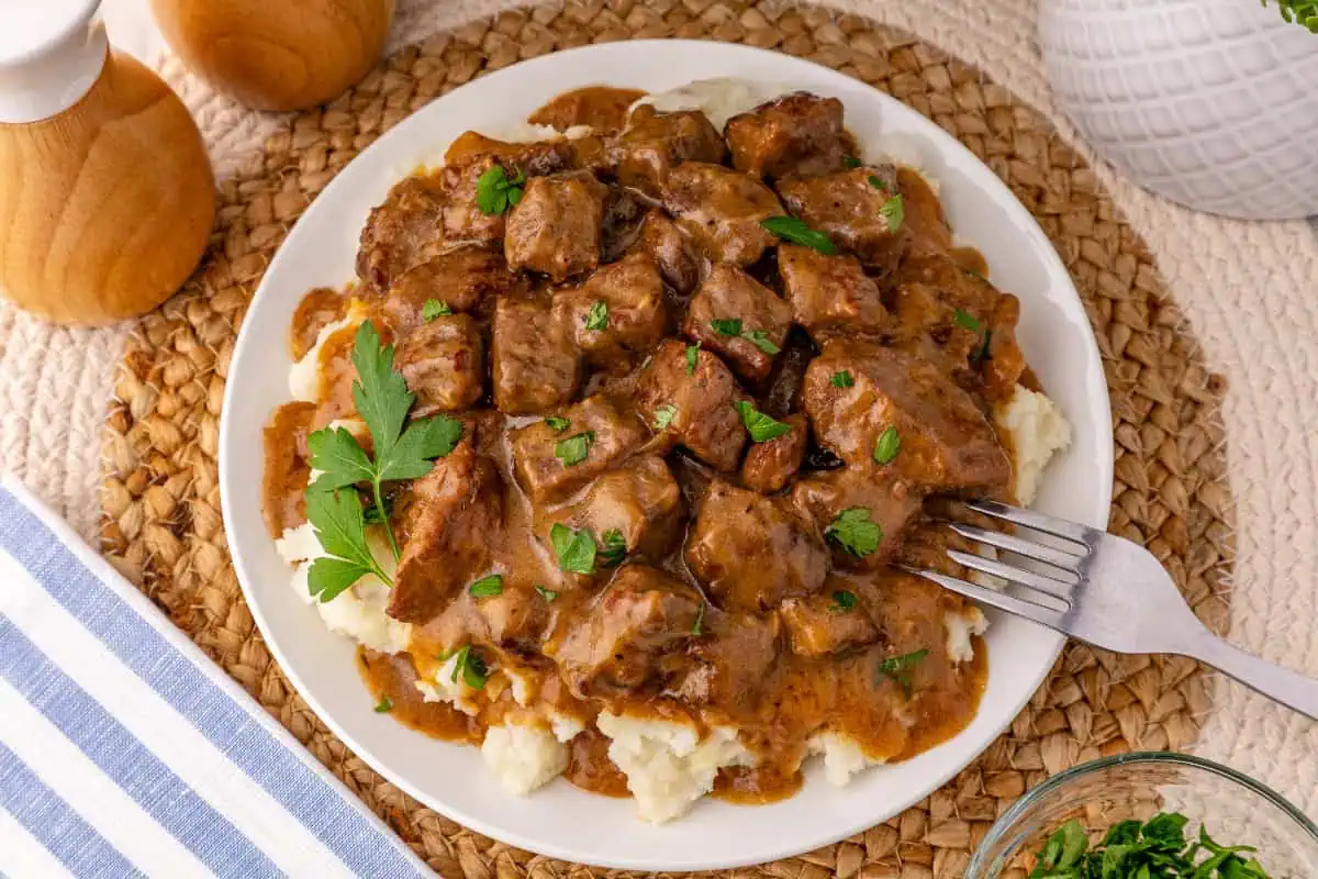 A plated serving of beef tips and gravy over mashed potatoes on a placemat with a fork