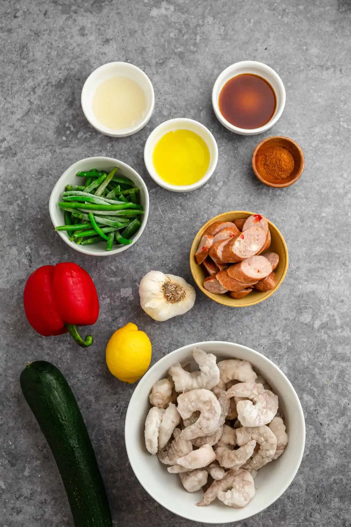 Jumbo shrimp, andouille sausage, zucchini, green beans, bell pepper, lemon, garlic bulb, and Cajun seasoning laid out for sheet pan Cajun shrimp and sausage