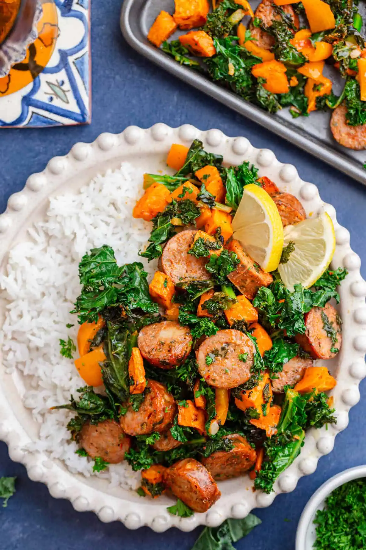 Sheet pan sausage, kale, and sweet potatoes roasted with crispy edges and lemon wedges on white bowl with rice