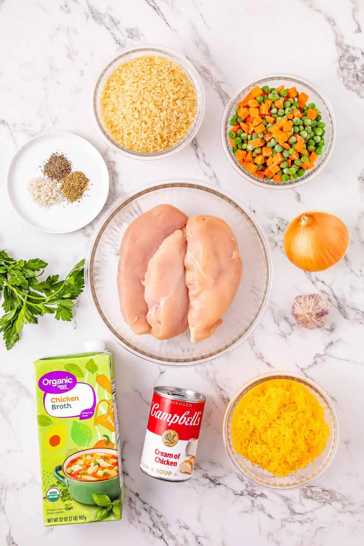Overhead view of parboiled rice, raw chicken breasts, chicken broth, cream of chicken soup, frozen peas and carrots, shredded cheddar, onion, and garlic for crockpot chicken and rice casserole