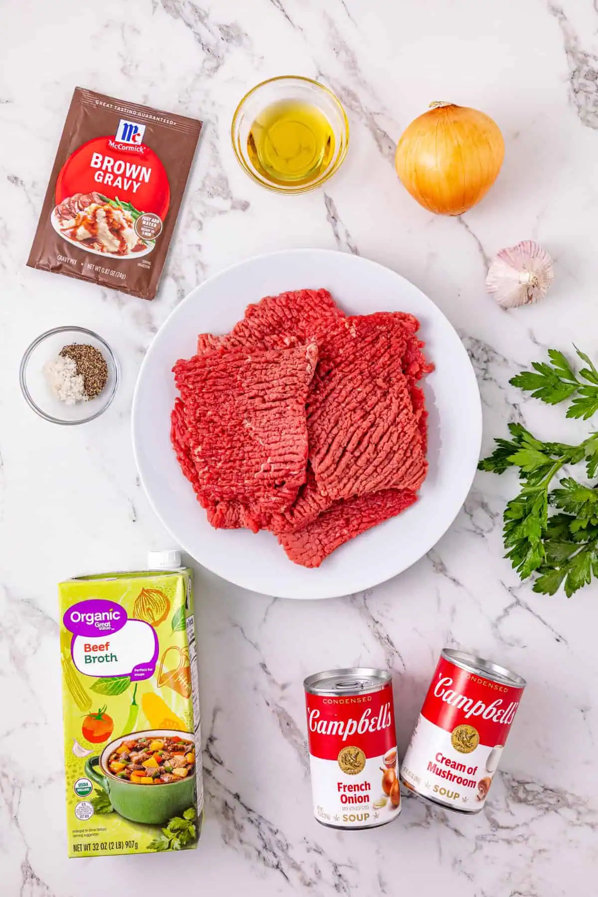 Overhead view of cube steaks, sliced onion, garlic, French onion soup, cream of mushroom soup, beef broth, and brown gravy mix for crockpot cube steak