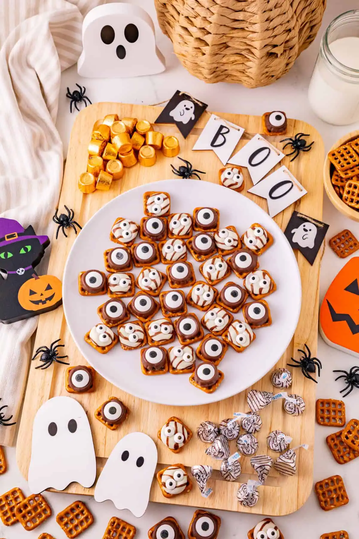 Halloween monster eye pretzels topped with Rolos, Hershey’s Hugs, and candy eyes on a white plate on a wooden cuutting board with ghosts and pumpkins