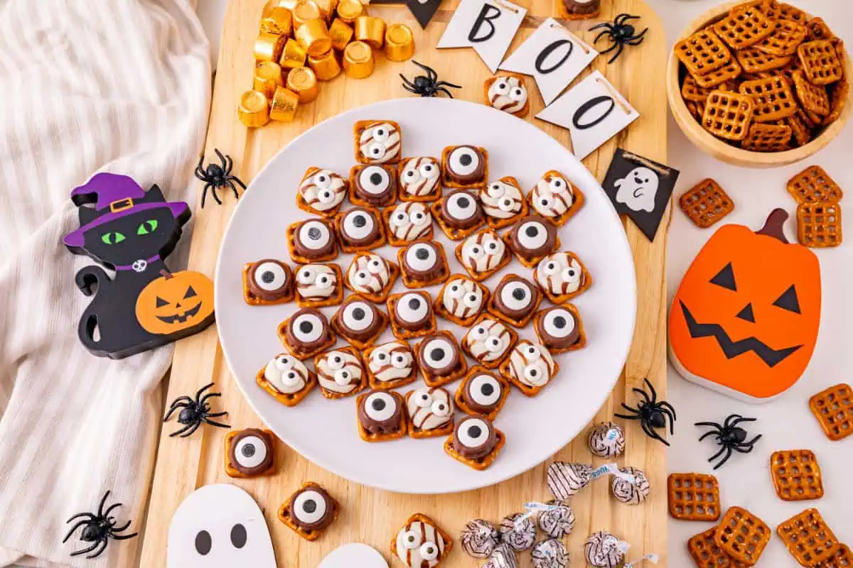 Halloween monster eye pretzels with one-eyed Rolo monsters and two-eyed Hershey’s Hug monsters on a white plate with a cutout wooden pumpkin, ghosts, and black cat sitting next to a pumpkin